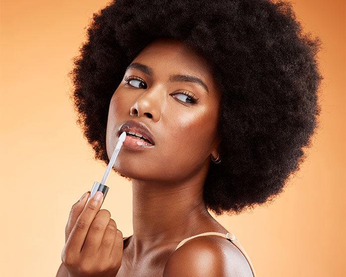Black woman, afro and natural beauty lip gloss treatment for a healthy, shiny and transparent tint, Cosmetics, apply and beautiful face of African model holding makeup tool at orange background