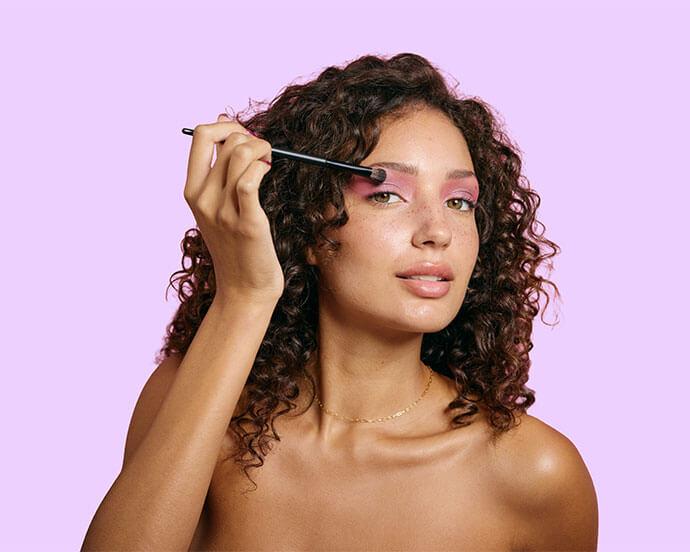 A woman with curly hair applies pink eyeshadow using a makeup brush against a pastel purple background