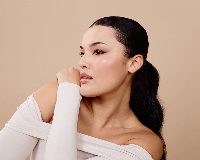A woman with sleek pulled-back hair poses in profile, showcasing soft, blended eyeliner and a natural glow against a neutral background