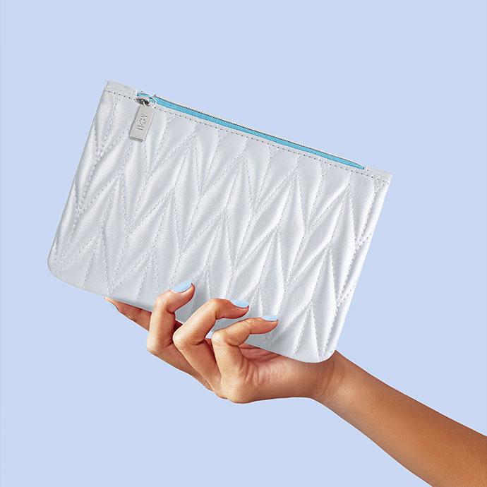 A hand holds a white quilted IPSY Glam Bag against a light blue background. The bag features a geometric stitched pattern, a silver IPSY zipper pull, and a contrasting light blue zipper.