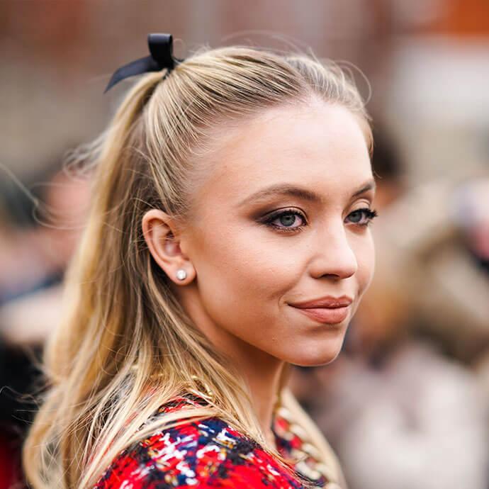 Actress Sydney Sweeney wears earrings, a ribbon on the hair, a red tweed jacket with golden chains