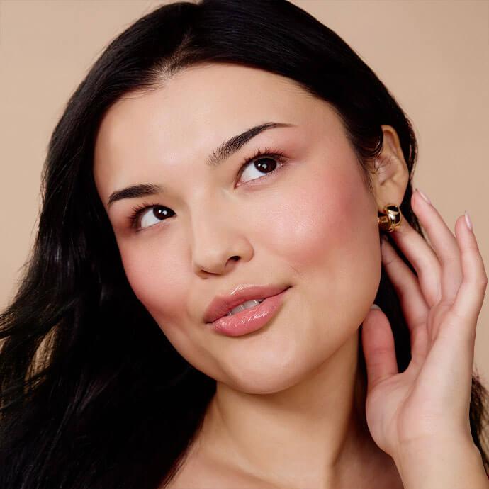A close-up portrait of a woman with smooth, glowing skin, dark hair, rosy cheeks, and glossy lips, touching her ear with a subtle, thoughtful expression