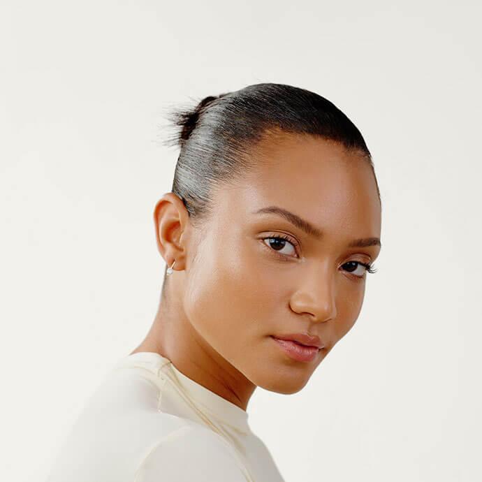 A close-up portrait of a woman with smooth skin and a sleek bun, wearing minimal makeup that enhances her natural glow with subtle foundation, light blush, and neutral lips
