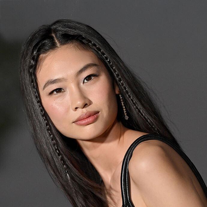 An image of a young woman with long black hair and subtle makeup gazes softly at the camera, wearing a black outfit and a small braided detail in her hair