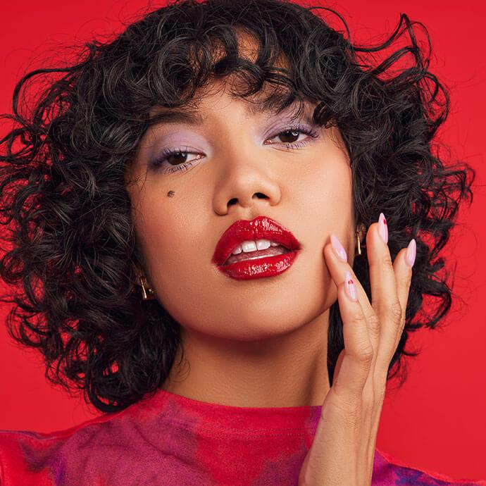 An image of a woman with curly short hair, bold red lips, and lavender eyeshadow poses against a vivid red backdrop, touching her face with polished nails