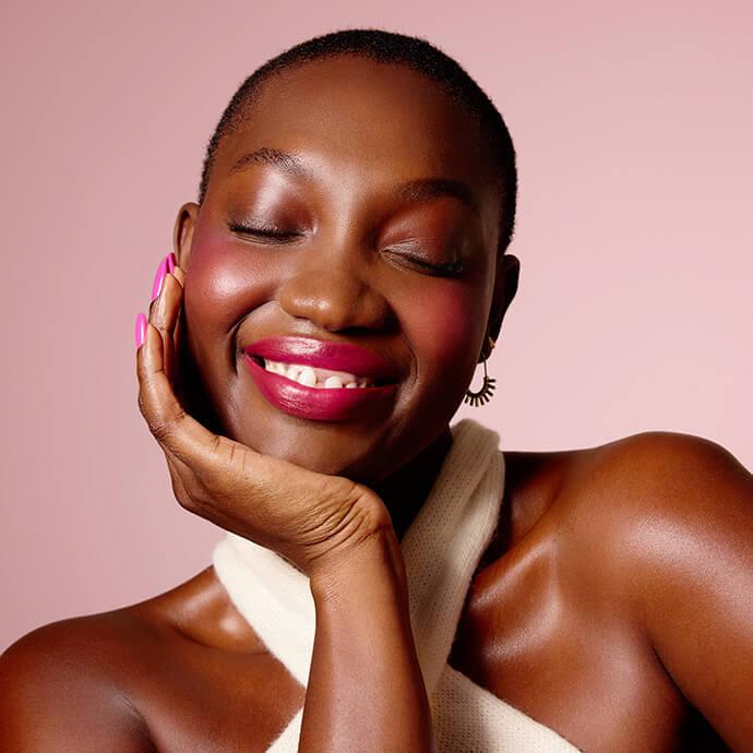 Smiling person with eyes closed and head resting on hand, wearing glossy pink lipstick, soft blush, and shimmery eye makeup against a blush-pink background.