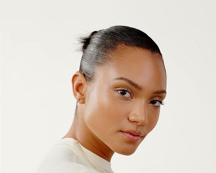 A close-up portrait of a woman with smooth skin and a sleek bun, wearing minimal makeup that enhances her natural glow with subtle foundation, light blush, and neutral lips