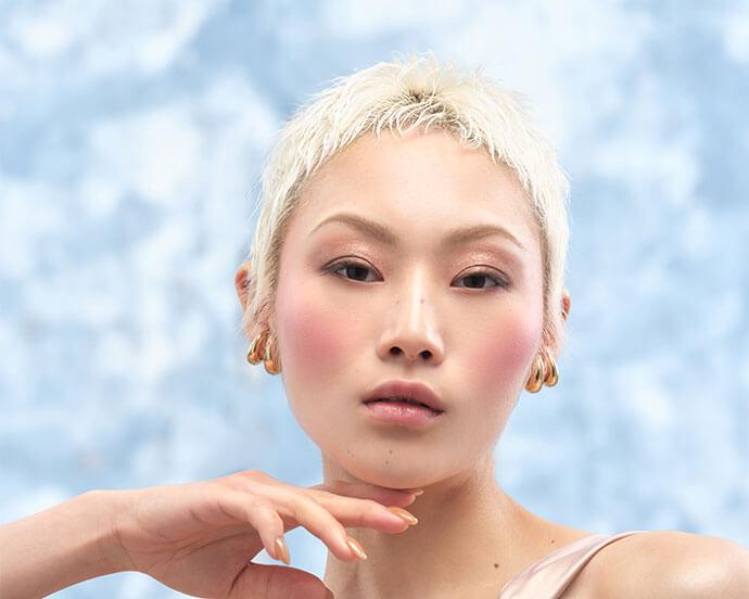 A close-up portrait of a woman with a short platinum blonde hairstyle and rosy makeup—soft pink blush, subtle pink eyeshadow, light eyeliner, and glossy nude lips—posing with a graceful hand against a blue backdrop