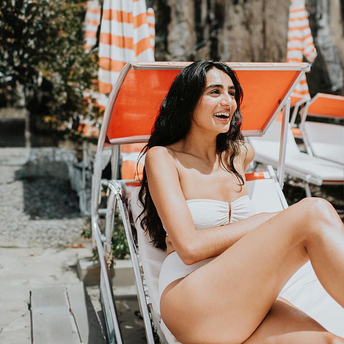 A beautiful Indian woman laughs, looking at something out of frame, as she reclines on a sun lounger within a holiday resort. She wears a white strapless bikini.