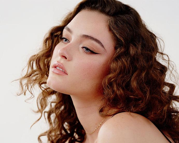 A portrait of a woman with curly brown hair and natural makeup—winged eyeliner, rosy cheeks, glossy nude lips, subtle highlighter, and neutral eyeshadow—gazing upward with a serene expression