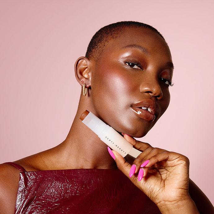 A model with a close-cropped haircut holds a Fenty Beauty foundation stick near her chin, showcasing luminous skin, glossy lips, and soft glam makeup against a pale pink background.