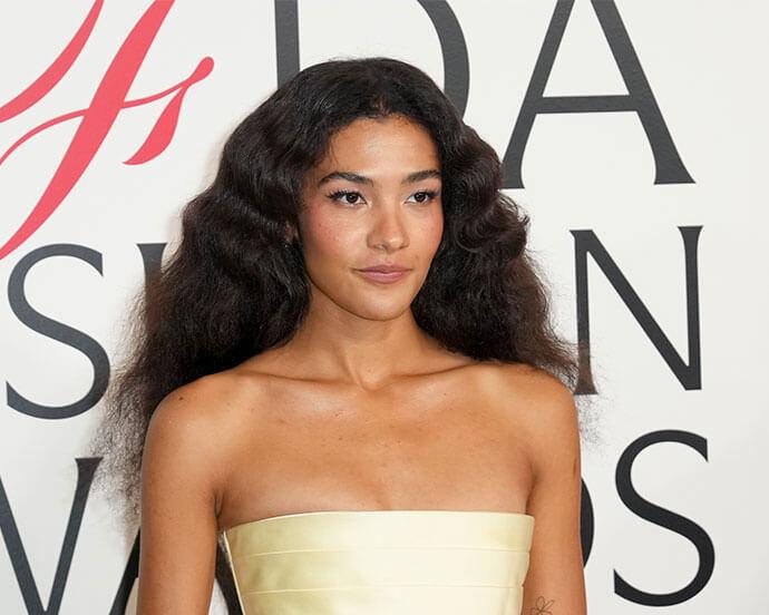 An image of Olivia Dean with voluminous, wavy black hair wears a strapless pale yellow dress at a red-carpet event, posing gracefully against a backdrop with bold lettering