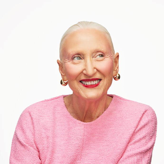A smiling, older woman with slicked-back white hair, wearing a textured pink top and gold hoop earrings, is looking off to the side against a bright white background