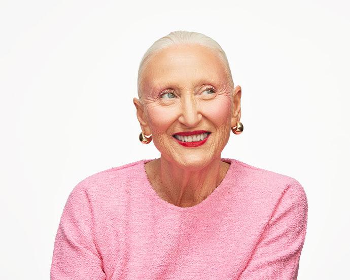 A smiling, older woman with slicked-back white hair, wearing a textured pink top and gold hoop earrings, is looking off to the side against a bright white background