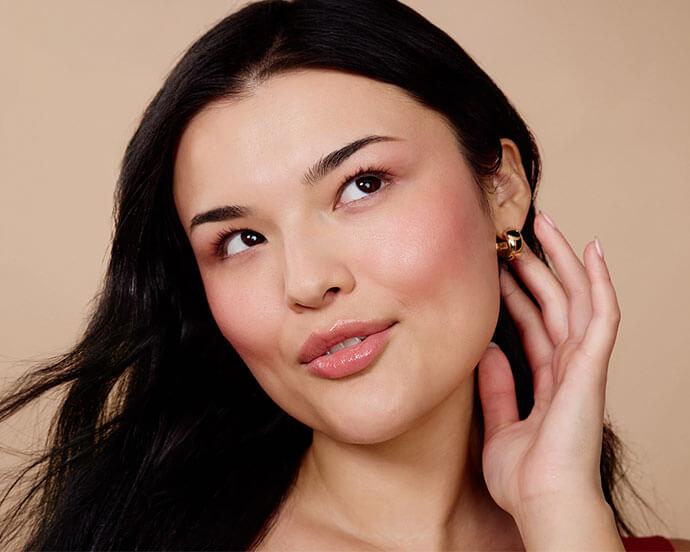 A close-up portrait of a woman with smooth, glowing skin, dark hair, rosy cheeks, and glossy lips, touching her ear with a subtle, thoughtful expression