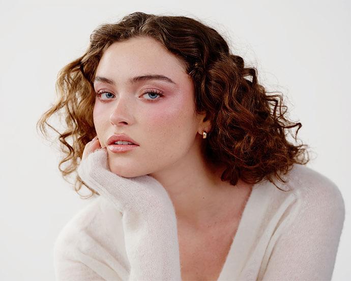 A close-up portrait of a person with short curly hair wearing soft sculpted makeup, including rosy blush, subtle contour, and glossy lips, resting their chin on their hand against a light neutral background.