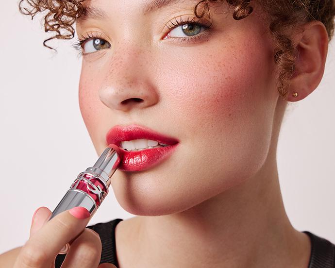 Close-up of model applying glossy red lipstick with softly diffused edges, illustrating the blurred lip trend with a modern, soft-focus finish