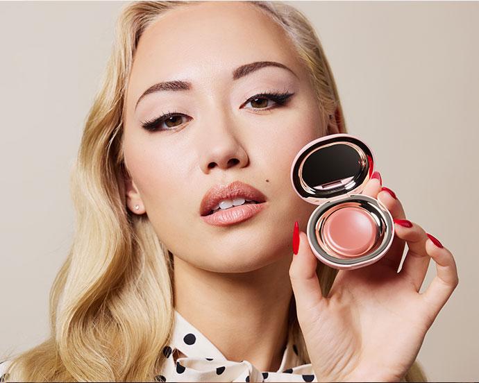 Close-up beauty portrait of a model demonstrating cream blush application, showcasing a soft radiant complexion and holding an open cream blush compact for a step-by-step makeup tutorial.