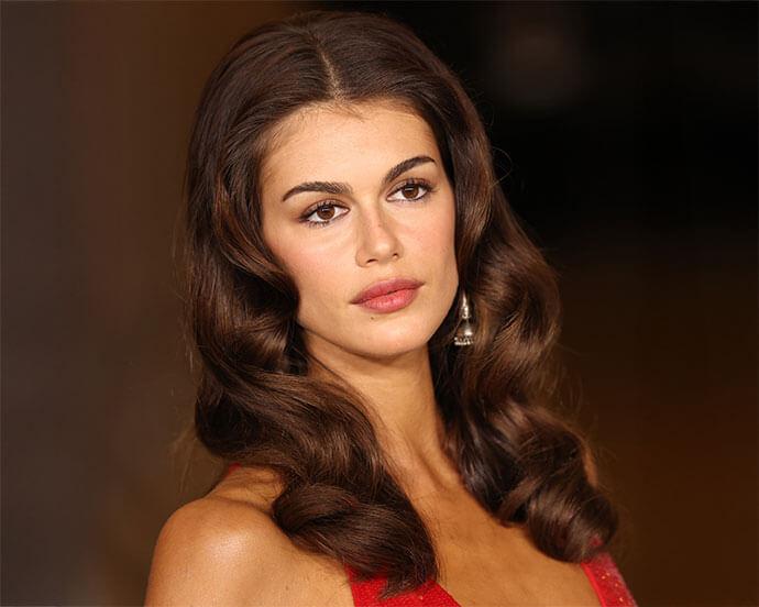 A stylish woman with voluminous, dark brown, center-parted waves and elegant makeup is captured in a headshot