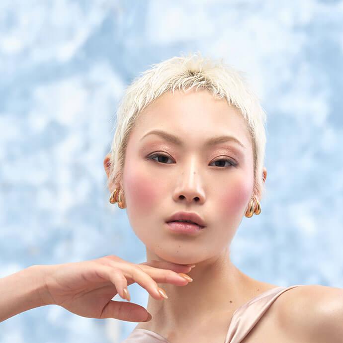 A close-up portrait of a woman with a short platinum blonde hairstyle and rosy makeup—soft pink blush, subtle pink eyeshadow, light eyeliner, and glossy nude lips—posing with a graceful hand against a blue backdrop