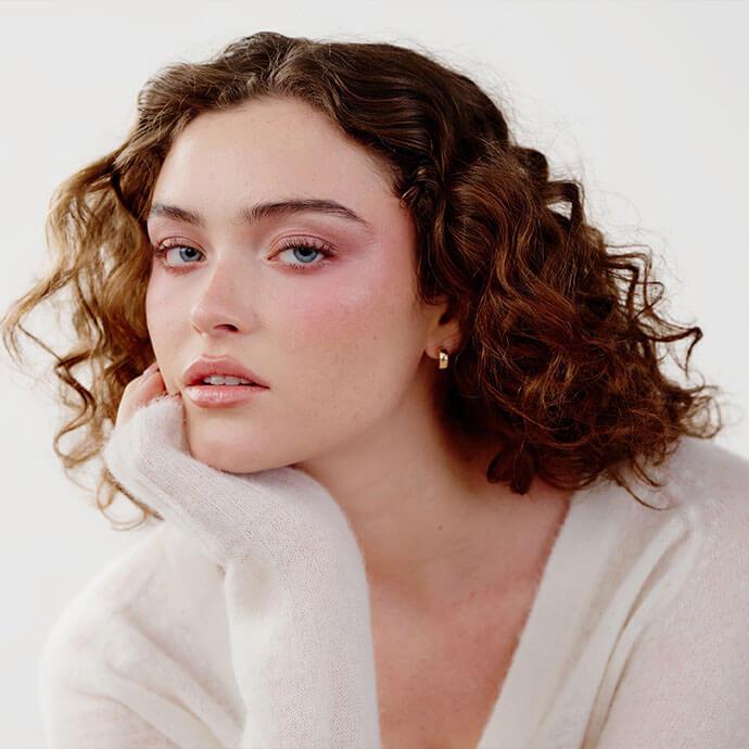 A close-up portrait of a person with short curly hair wearing soft sculpted makeup, including rosy blush, subtle contour, and glossy lips, resting their chin on their hand against a light neutral background.
