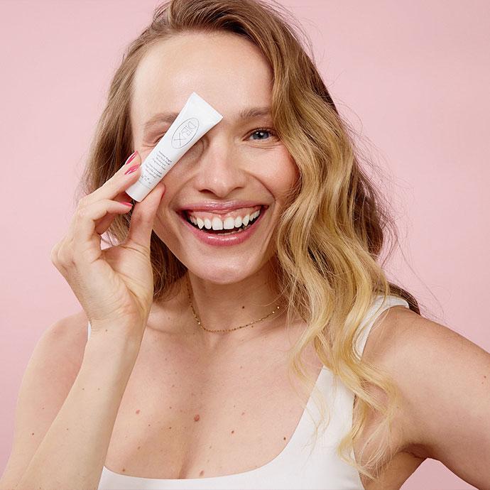 Smiling woman holds a DIEUX SKIN product tube near her eye against a pink background.