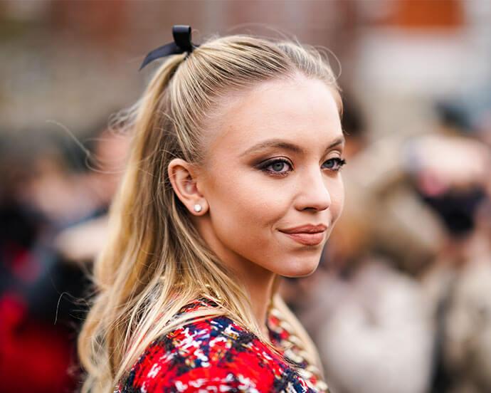 Actress Sydney Sweeney wears earrings, a ribbon on the hair, a red tweed jacket with golden chains