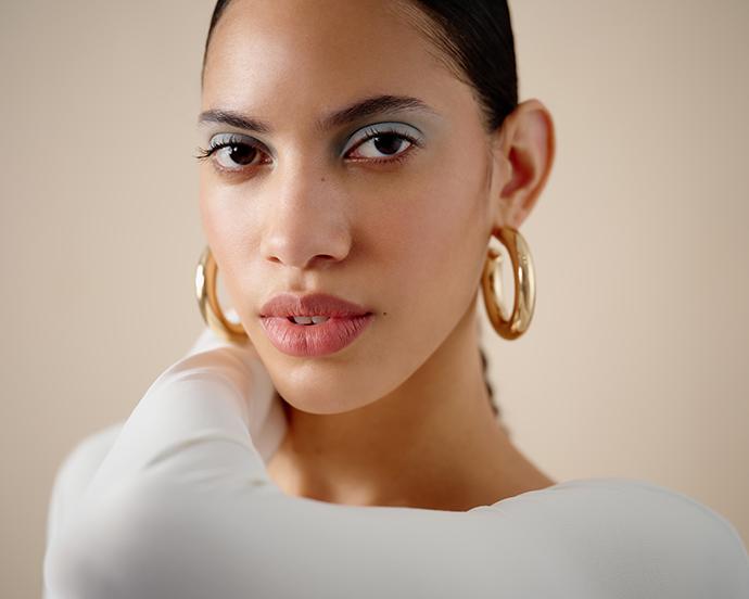 Model with slicked-back hair wearing frosted blue eyeshadow, glowing skin, and glossy lips for a modern soft glam makeup look