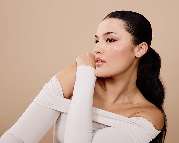 Model wearing a sleek low ponytail and soft, glowing spring makeup with rosy blush, glossy lips, and neutral eyeshadow against a beige background.