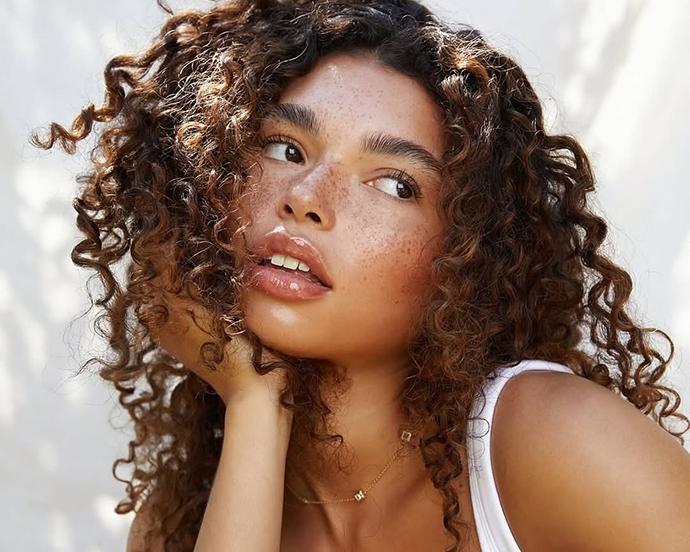 Model with caramelized brunette curly hair, featuring warm brown tones, natural texture, and sunlit dimension for a soft, glowy finish.