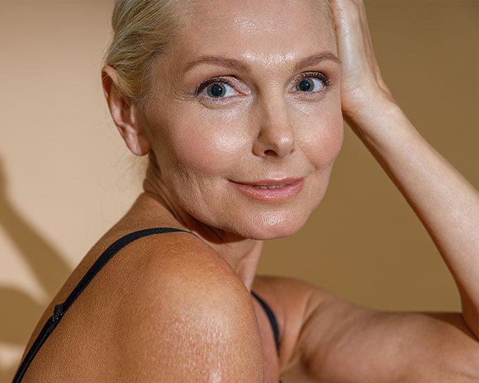 A middle-aged woman with fair skin and blonde hair, wearing a black strap top, looks directly at the camera with a gentle smile while resting one hand on her head.