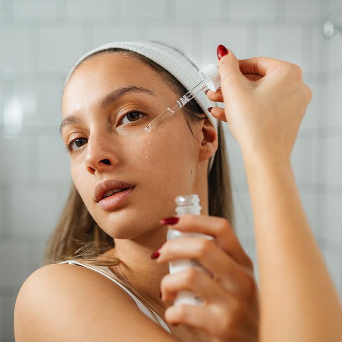 A woman with a white headband and red nail polish applies a clear facial serum to her cheek using a glass dropper in a bathroom