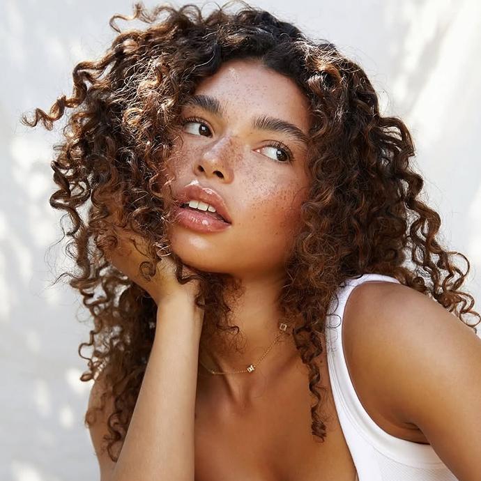 Model with caramelized brunette curly hair, featuring warm brown tones, natural texture, and sunlit dimension for a soft, glowy finish.