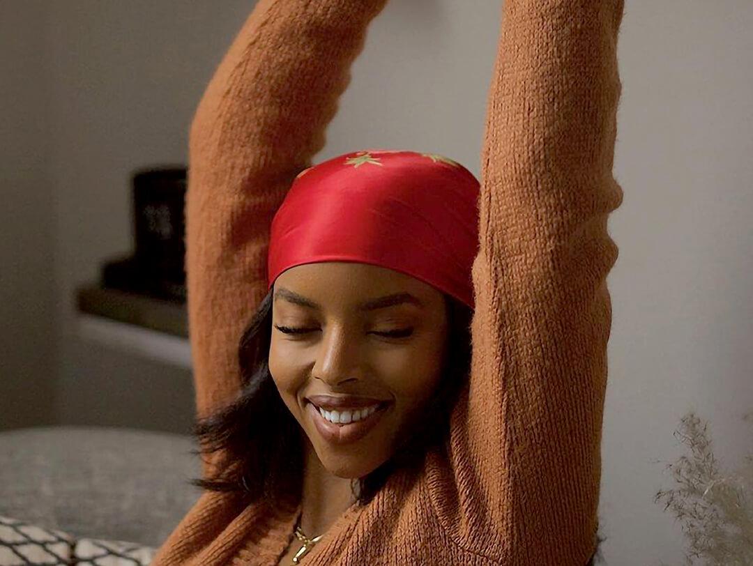 Smiling Aysha Sow rocking an orange bandana and posing with her arms up in her living room Desktop