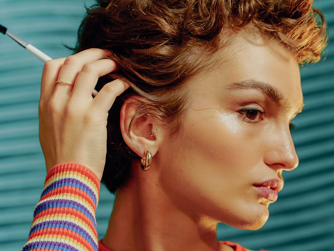 Woman with short hair and a natural makeup look touching her hair with a spoolie in hand Desktop