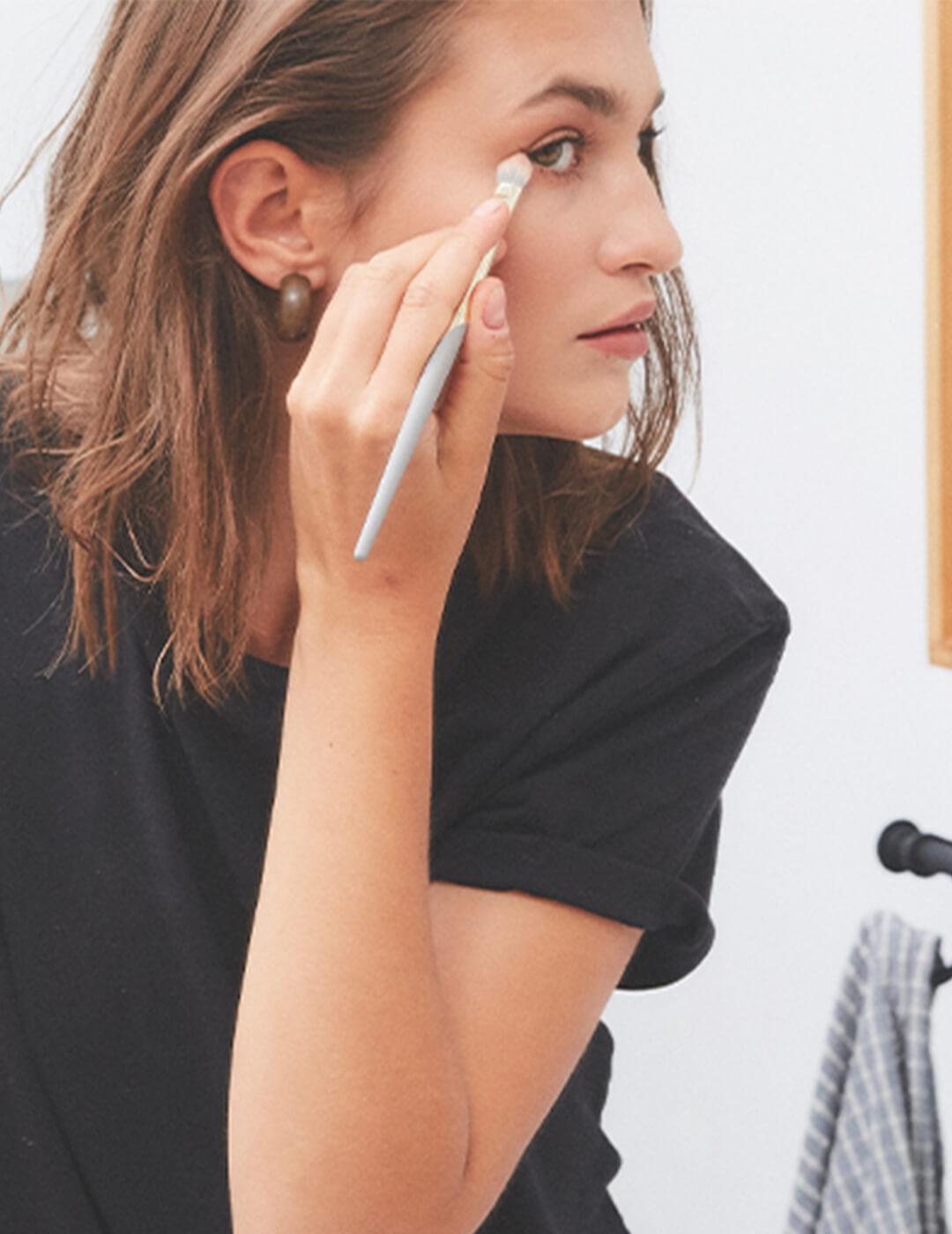 An image of a woman looking at the mirror while applying cosmetics in her eye area Mobile