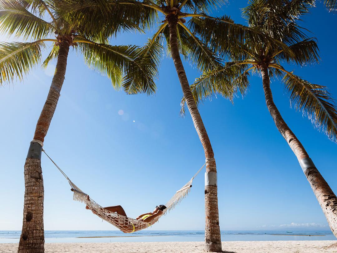 A photo of model relaxing on a sunny beach in a hammock between two coconut trees Desktop