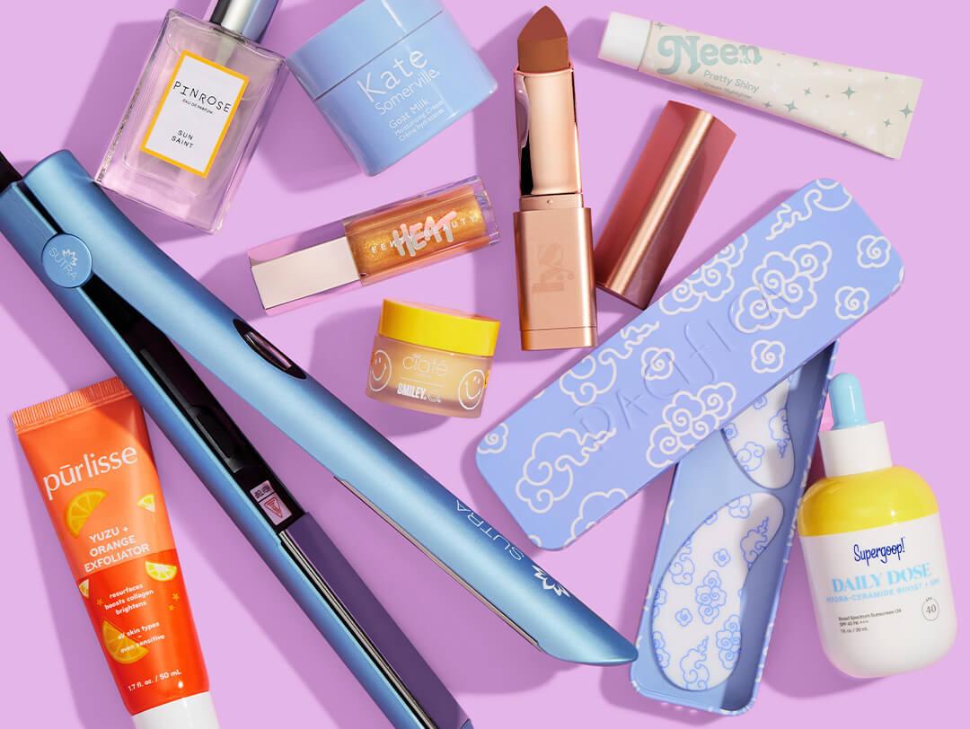 Skincare, makeup, and hair products and tools from various brands scattered on lavender background Desktop