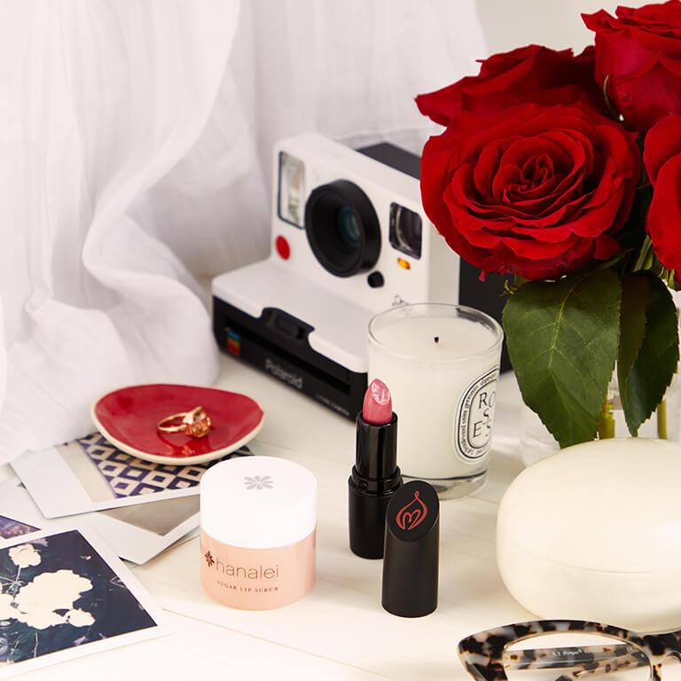 A flatlay image of lip products, polaroid camera, roses, candles, and a ring in a saucer and a closeup image of a model applying a bold red lipstick Mobile