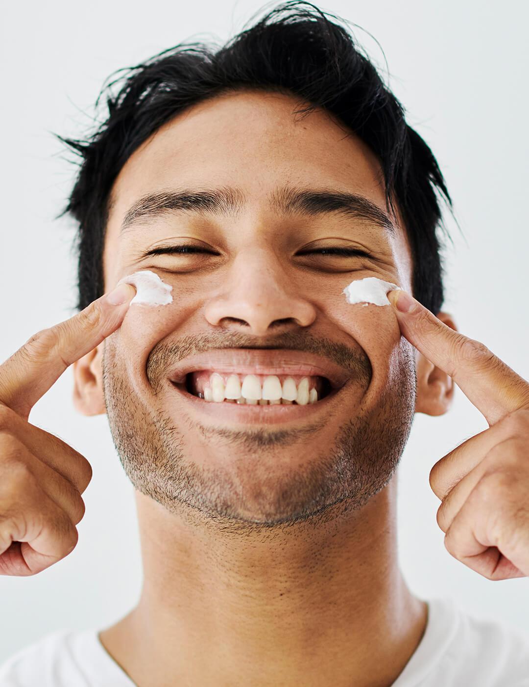 Man grooming, applying face cream or lotion, enjoying self care against grey background with copy space. Guy smiling while doing treatment and skincare. Male trying an anti aging or hydrating facial Desktop