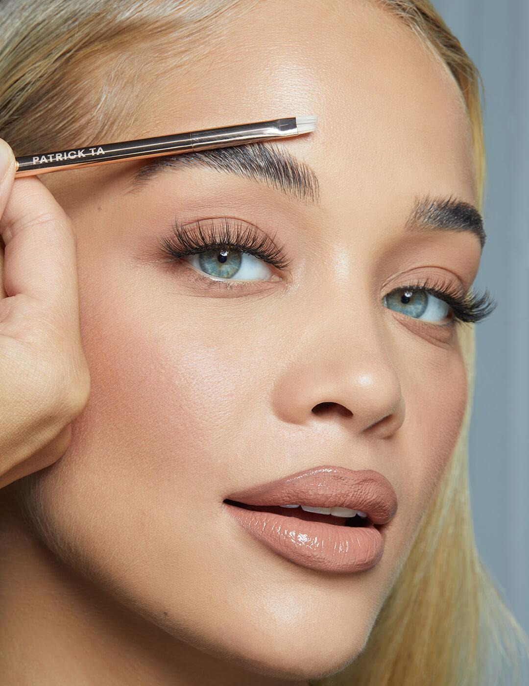 Close-up image of Jasmine Sanders with a neutral makeup look and a makeup brush touching her eyebrow Mobile