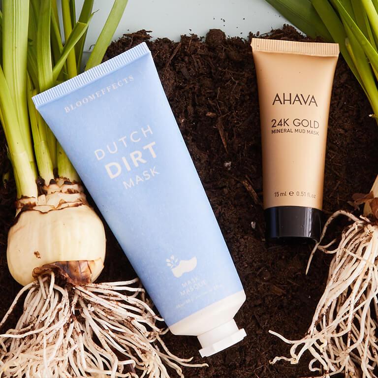 A flatlay image of BLOOMEFFECTS Dutch Dirt Mask and AHAVA 24K Gold Mineral Mud Mask and orchid bulbs on dirt soil Desktop