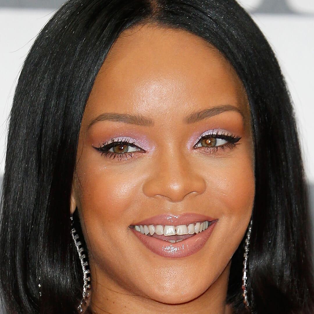 Close-up image of a smiling Rihanna wearing a shimmery lavender eyeshadow makeup look and silver dangling earrings Mobile