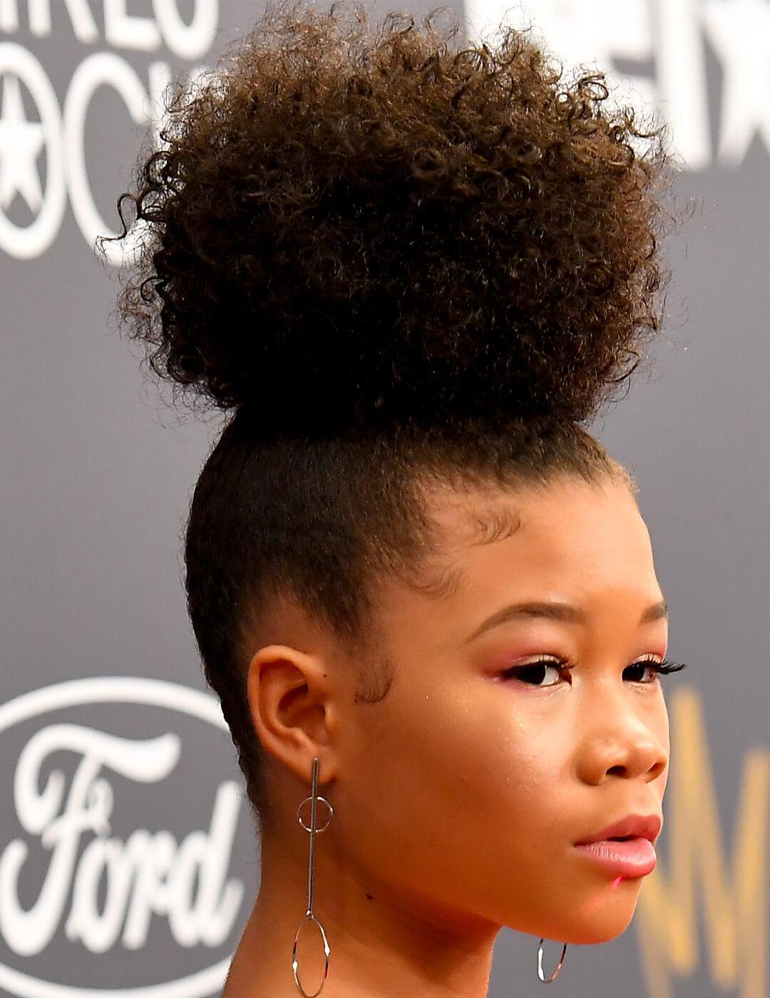 Storm Reid sporting a curly updo hairstyle and gold dangling earrings Mobile