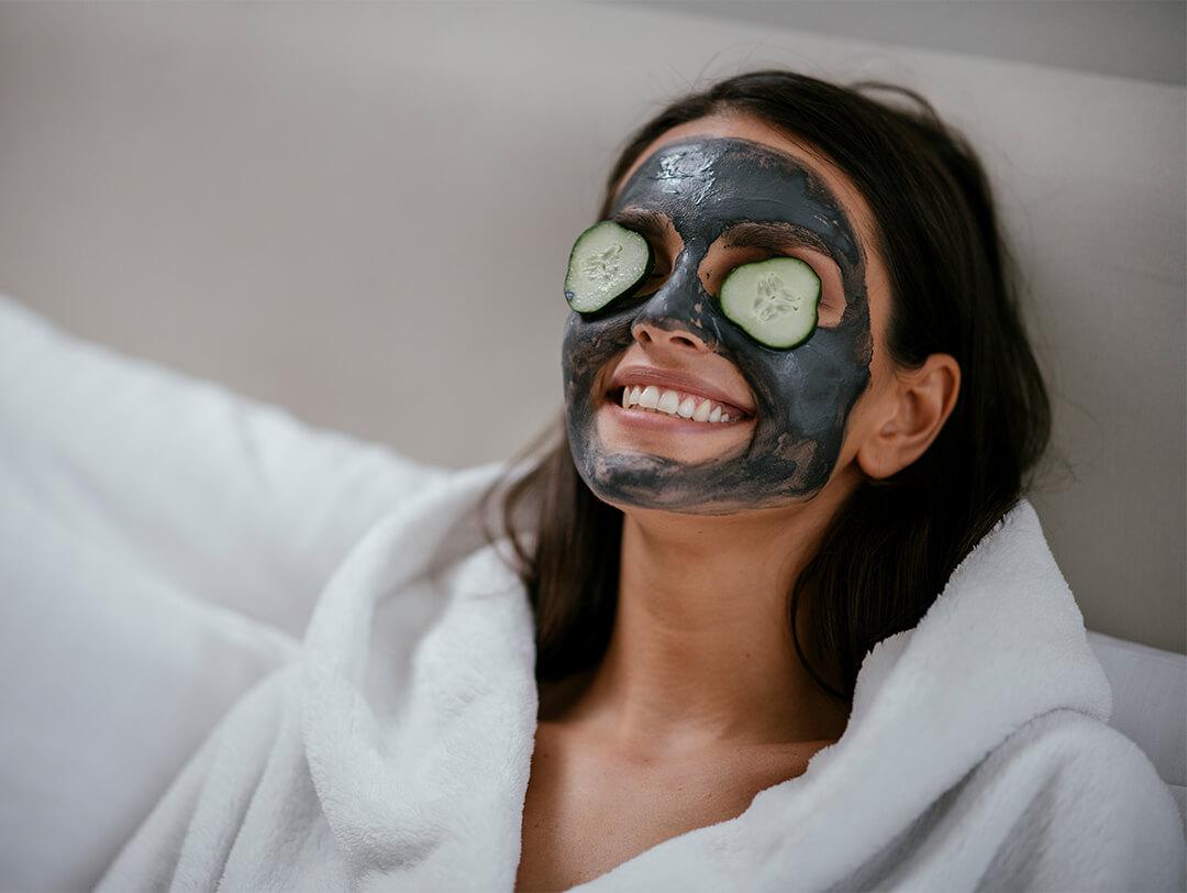 Image of a woman in a white robe with charcoal mask on her face and cucumber slices on her eyes Desktop