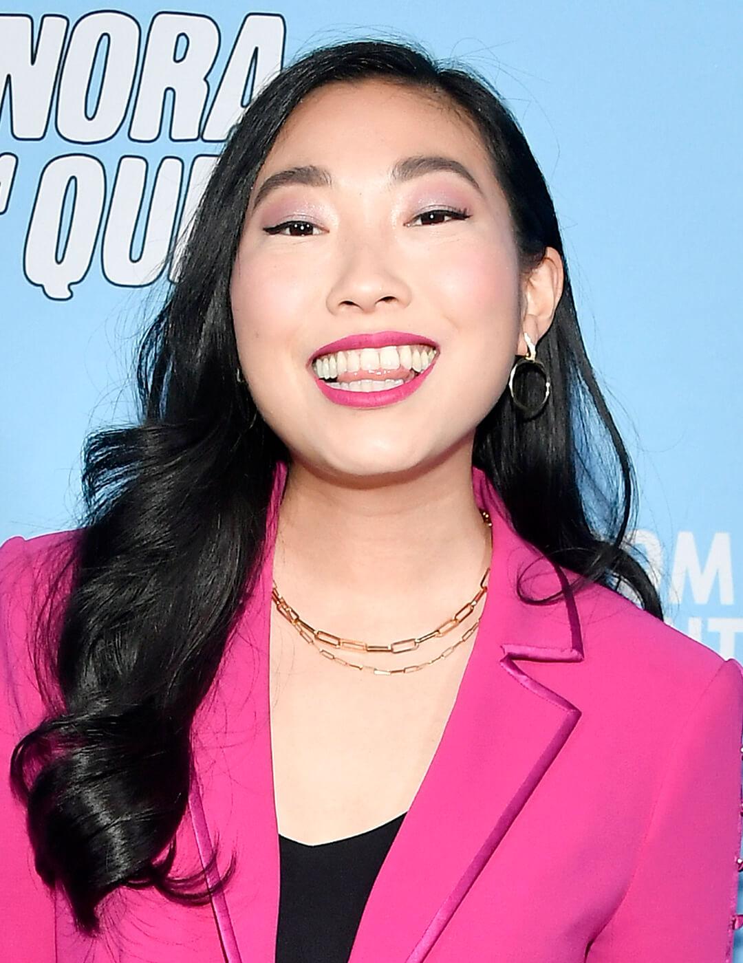 Cheerful Awkwafina rocking a pink suit, black v-nect top, gold necklaces, and wavy hairstyle on the red carpet Desktop