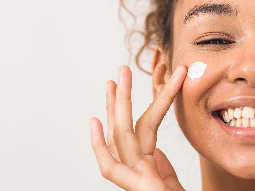 Close-up image of a smiling woman dotting face cream on her cheek Desktop