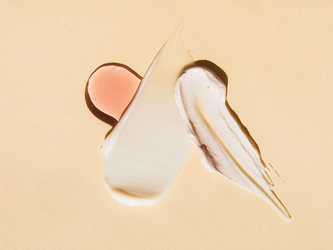 Swatches of textured and multi colored face creams and serum on a beige backdrop Desktop