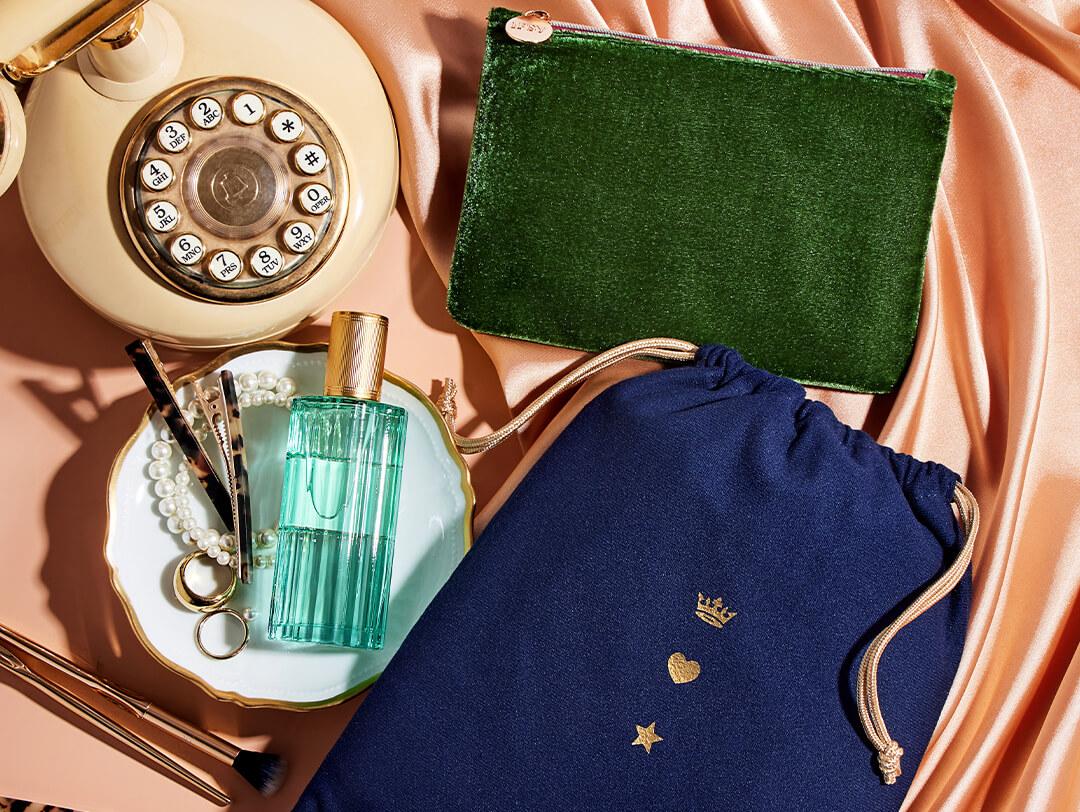 Flatlay image of November 2021 IPSY Glam Bag and Glam Bag Plus bags, vintage telephone, green bottle of perfume, gold and pearl accessories, on champagne-colored satin cloth Desktop