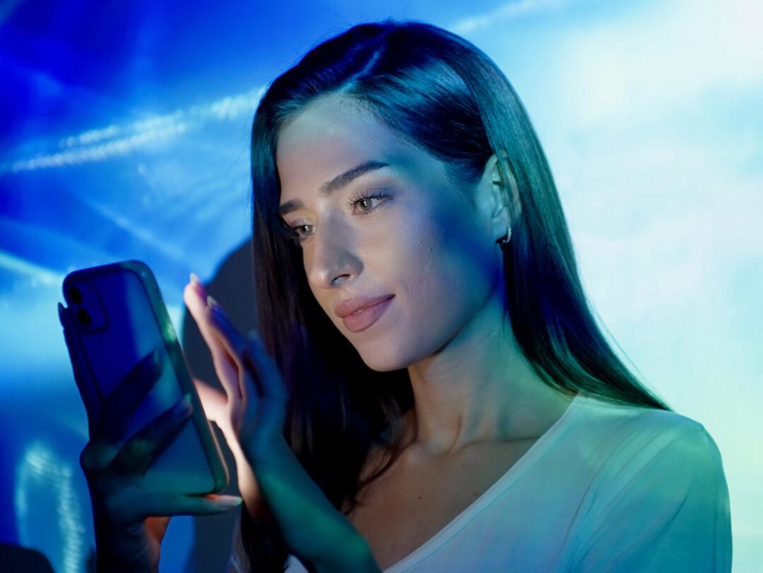 An image of a woman with a subtle smile, glancing at her phone wearing light makeup in a nude lipstick, dressed in a white t-shirt within a room illuminated by various shades of blue Desktop